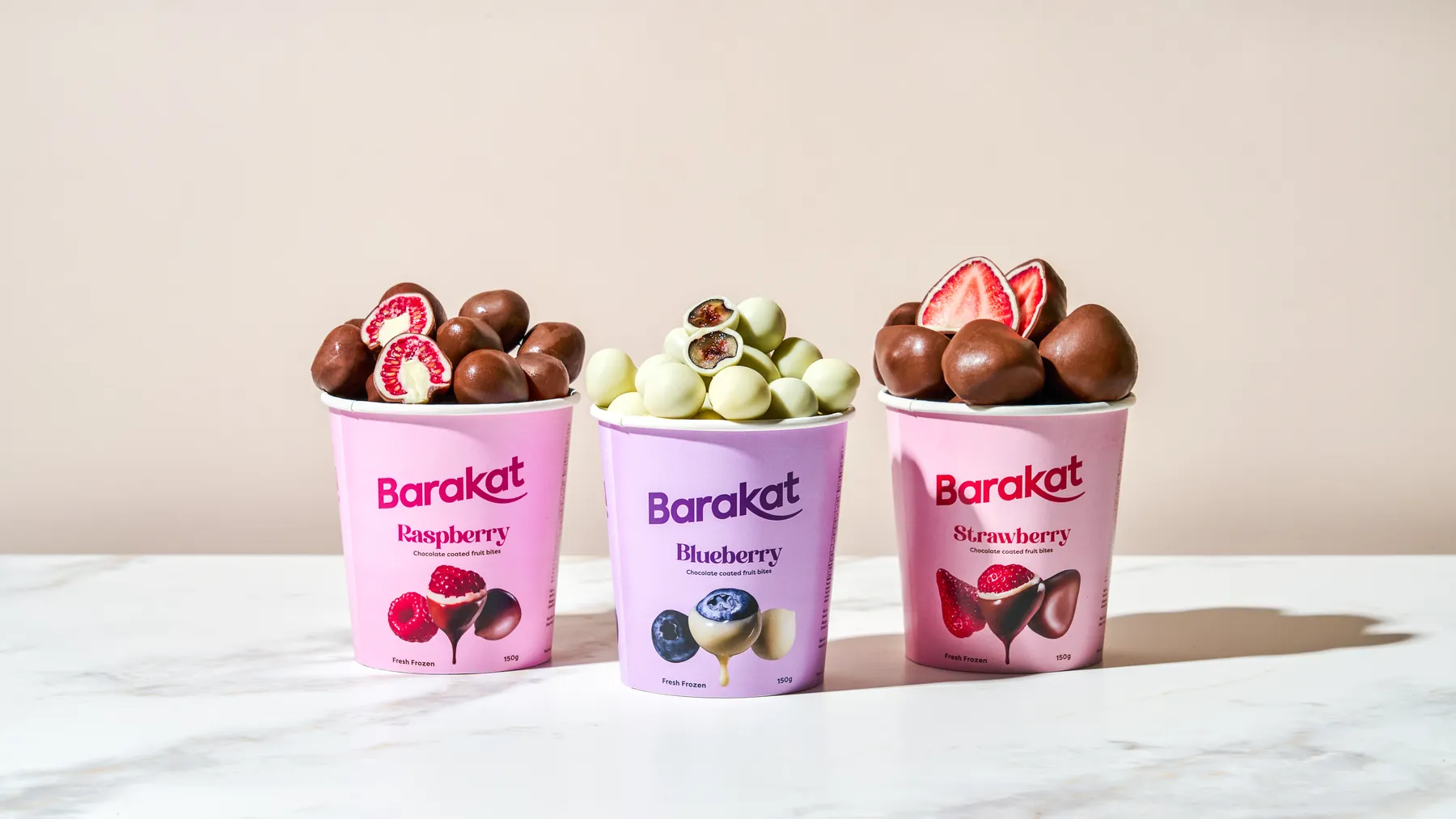 Three Barakat chocolate-coated fruit bite cups in pink and lilac on a marble surface against a warm blush backdrop